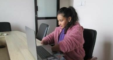 Image of a young woman (age 20) working on a laptop. The woman is a double amputee - both forearms end just below the elbow. She wears a purple shirt and a pink sweater.