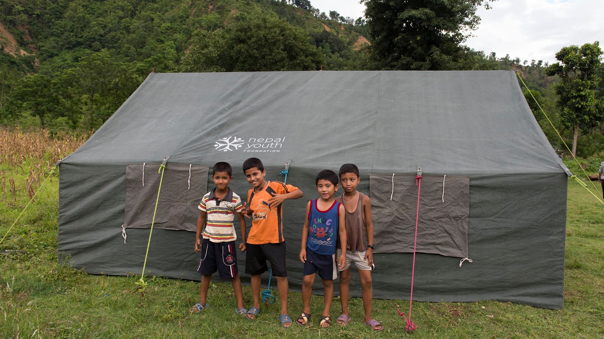 Four young boys stand smiling in front of a 20-person tent emblazoned with the NYF logo, green grass underfoot.