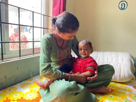 A young Nepali woman in a green dress sits cross-legged on a yellow floral bedspread, cradling her smiling toddler. A sunny window behind them allows light into the room.