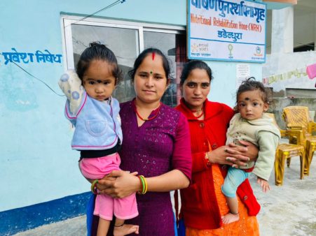 Two Nepali women (one in purple, one in red) stand side-by-side in front of the Dadeldhura Nutritional Rehabilitation Home. Each woman holds a toddler in her arms. The children have black dots (or kala tika) between their eyebrows and look curiously at the camera.